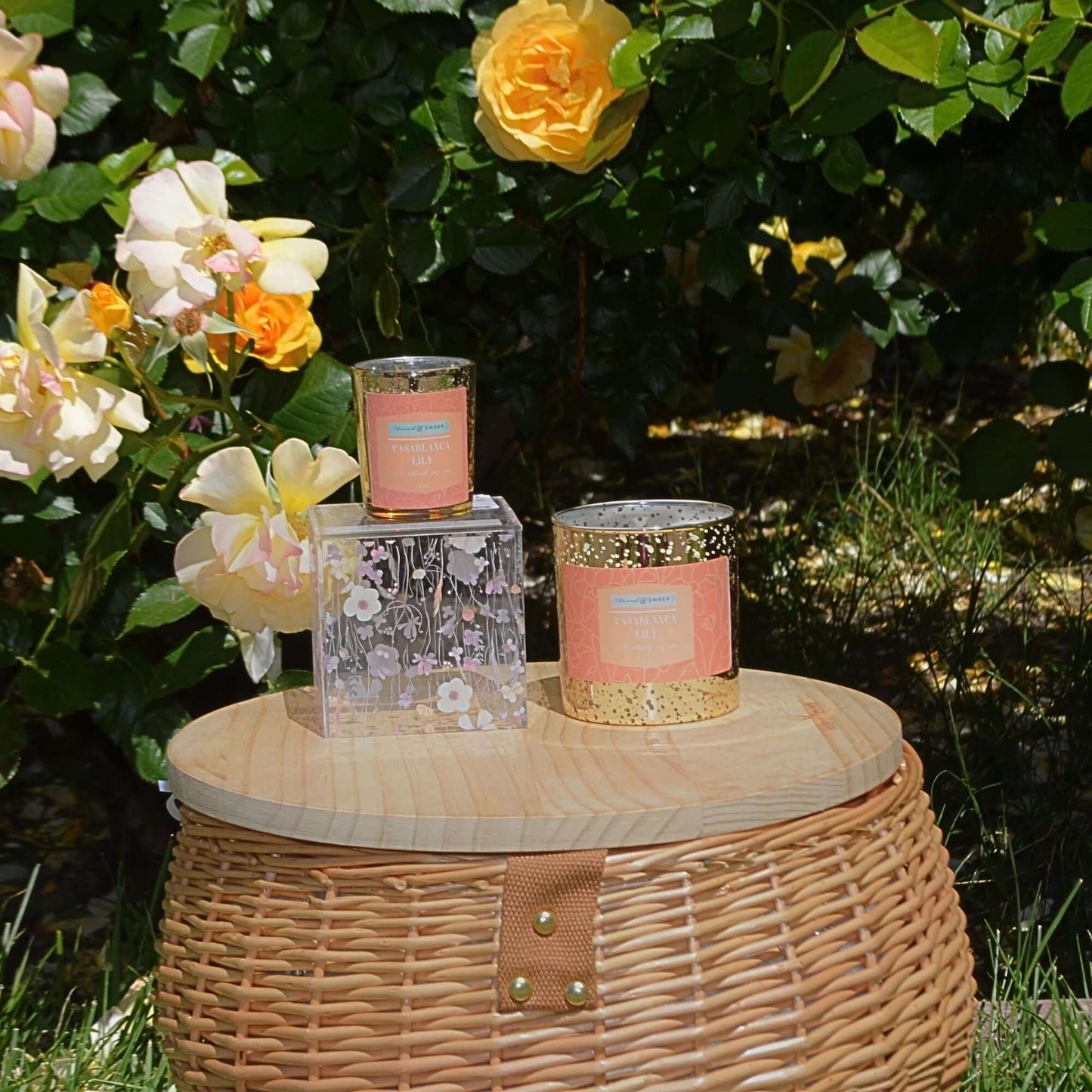 Casablanca Lily and Endless Escape candles displayed outdoors on a wooden surface surrounded by roses.
