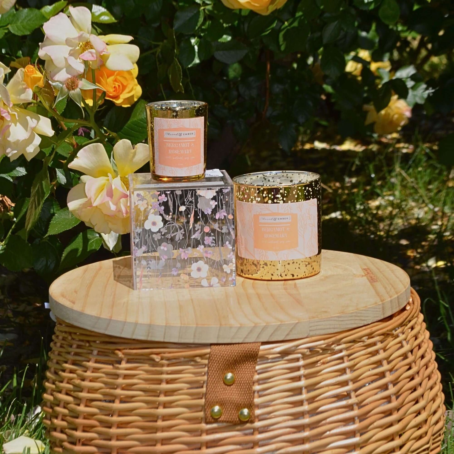 Bergamot & Rosemary candle in a gold mercury jar styled on a picnic basket with yellow roses in the background.