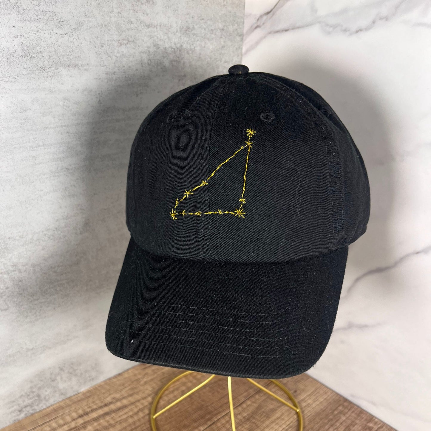 Front view of a black baseball hat with gold Cancer constellation embroidery