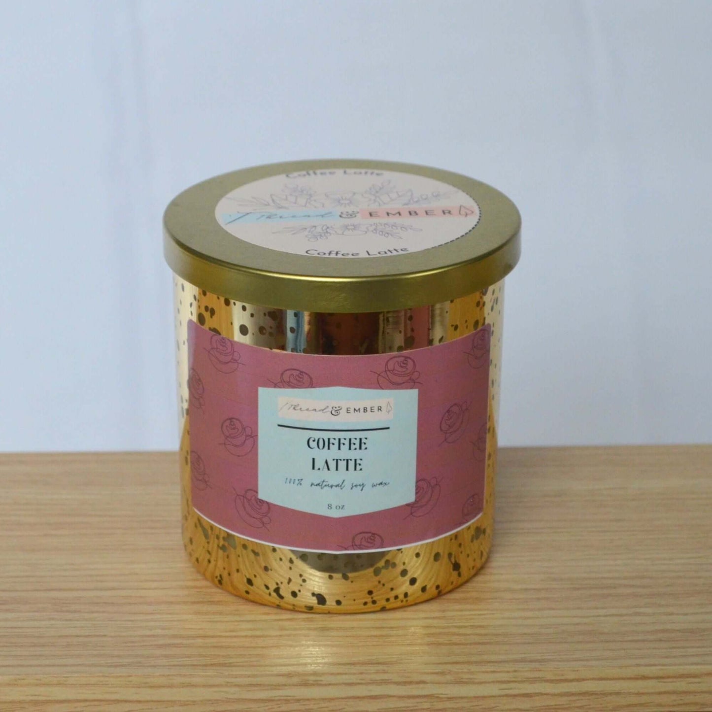 Coffee Latte candle with its gold lid on, featuring a pink label design, displayed on a wooden surface.