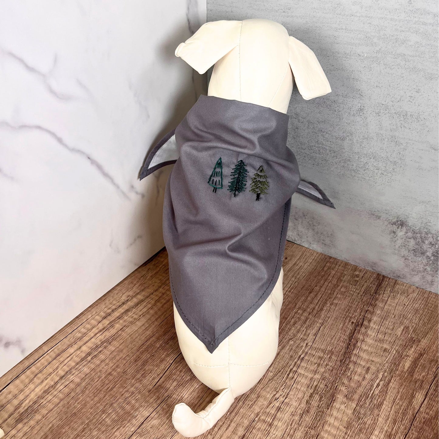 Gray pet bandana featuring hand-embroidered evergreen trees, modeled on a dog against a wooden surface background.