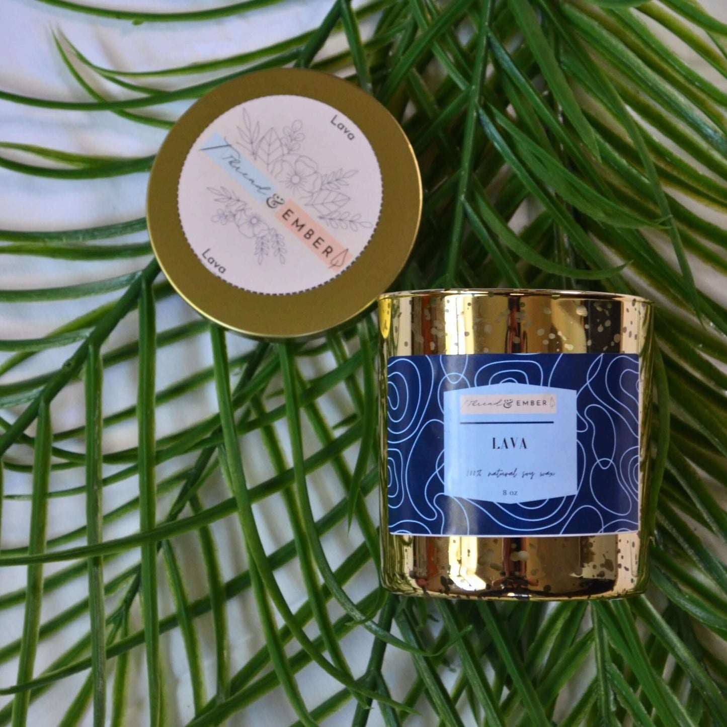 Thread & Ember Lava candle on tropical palm leaves, highlighting its golden speckled design and bold blue abstract label for an exotic ambiance.
