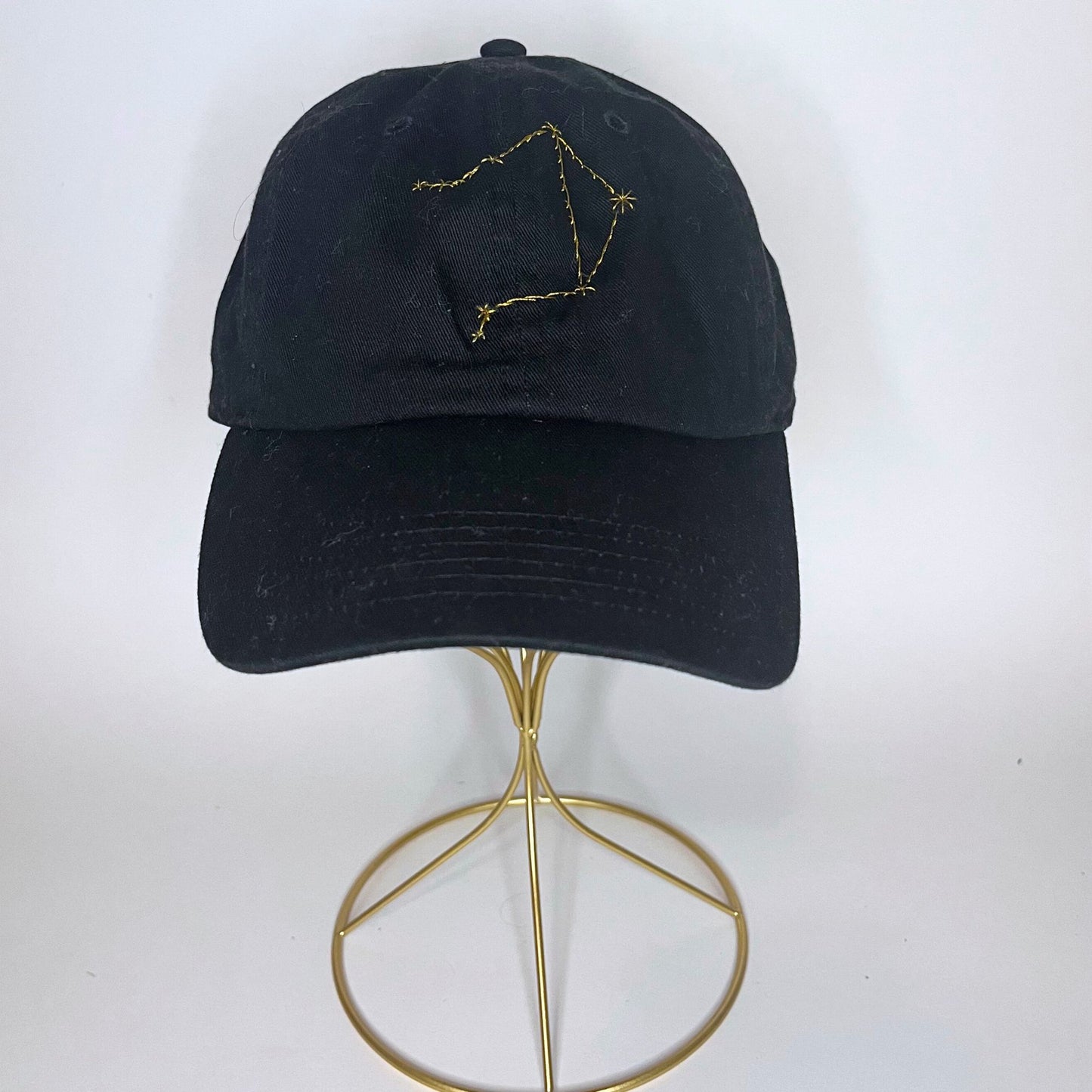 Front view of a black baseball cap featuring a yellow embroidered Libra zodiac constellation design.