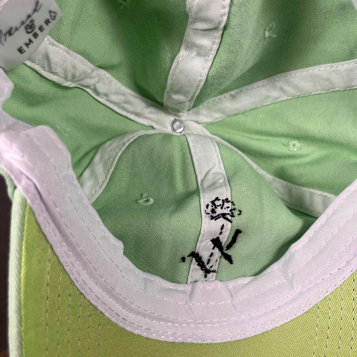 Inside view of a mint green cap with a white interior and an embroidered black poppy flower design on the inside panel.