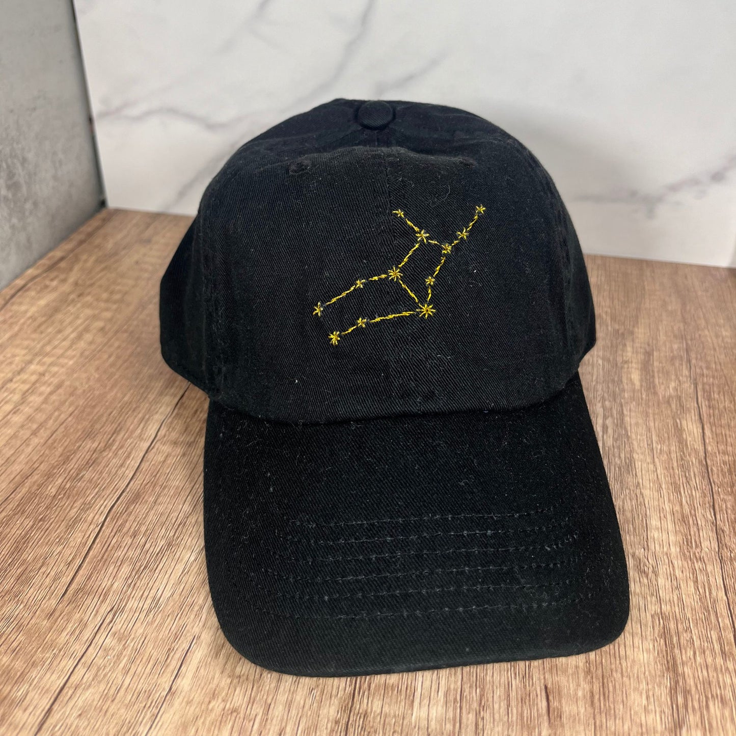Front view of a Virgo constellation black cap with intricate golden embroidery, placed on a minimalist stand.