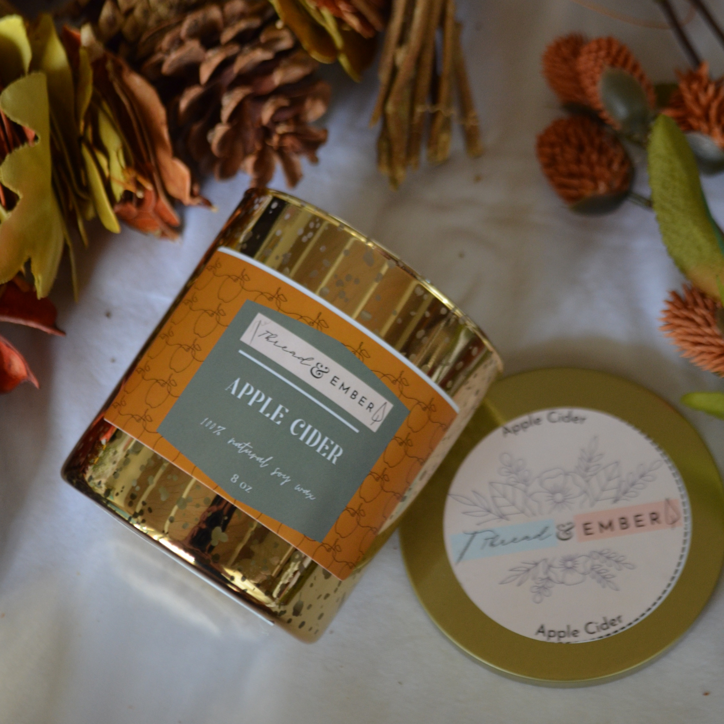 Thread & Ember Apple Cider Candle surrounded by autumn-themed decorations, showcasing its gold metallic jar and natural soy wax.