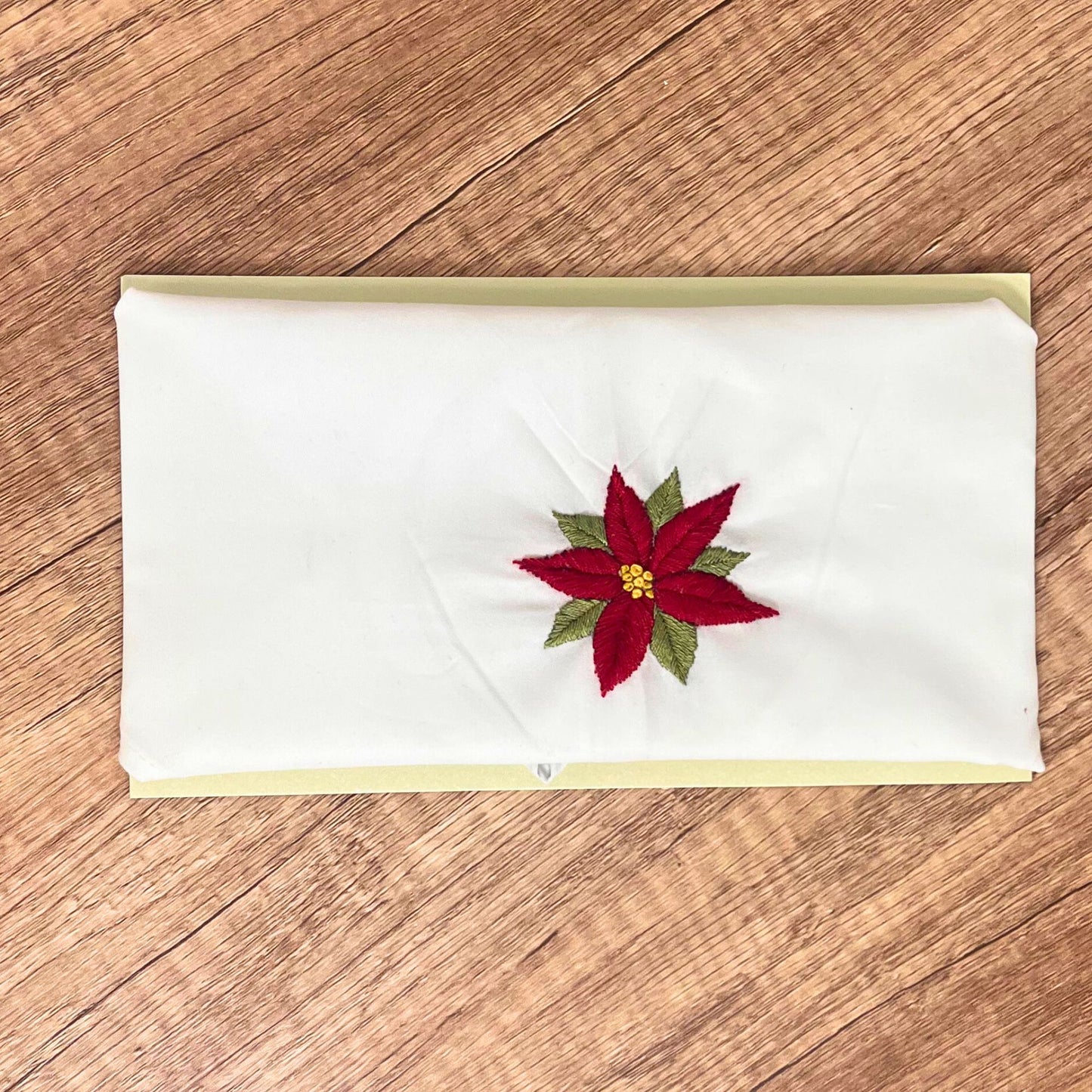 White cotton bandana featuring a hand-embroidered red poinsettia, displayed flat on a wooden surface.